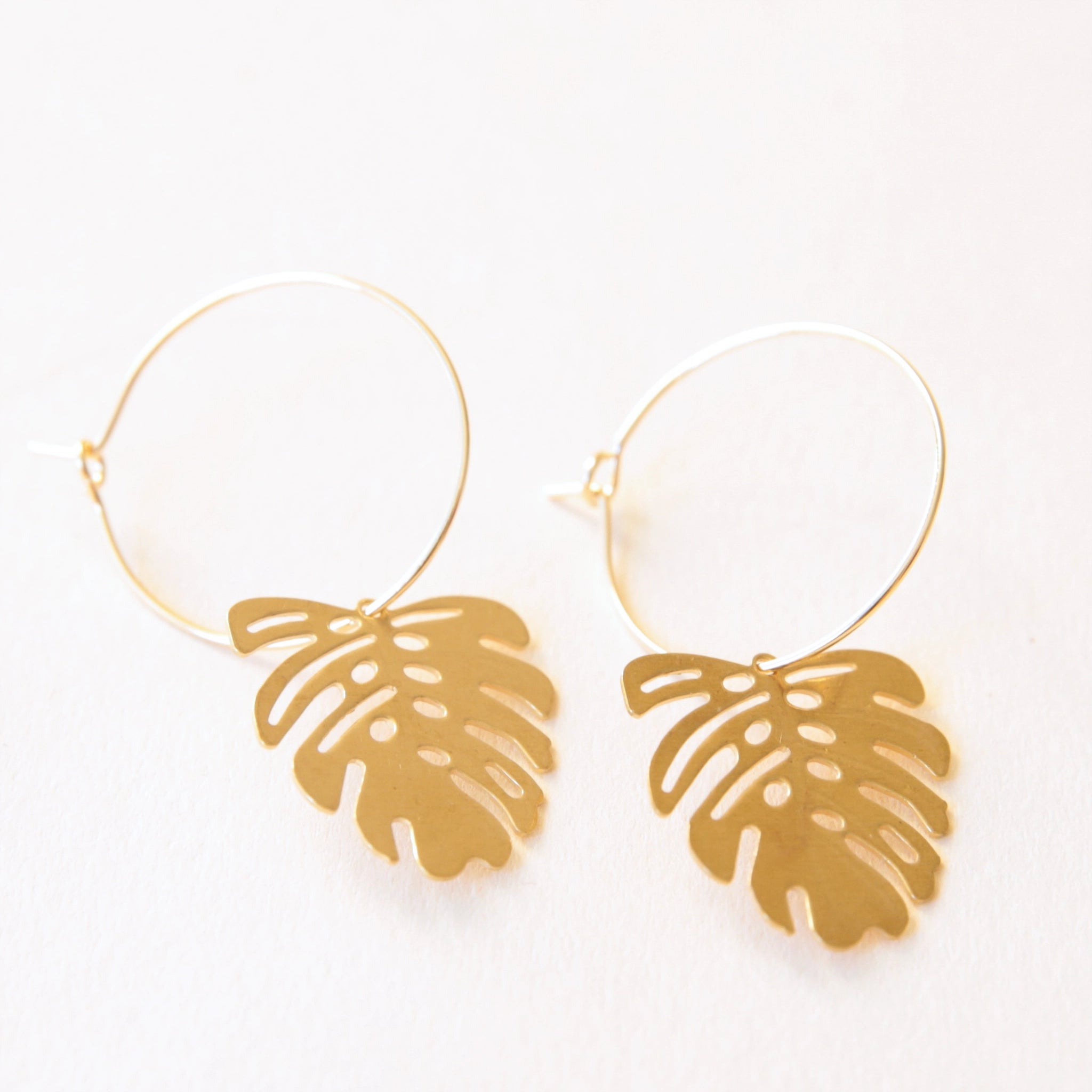 A thin gold hoop with a outlined gold monstera leaf charm hanging from the hoop.