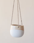 The smaller neutral ceramic hanging planter with rounded sides and a flat bottom along with a half cream half tan design and three jute strings as the hangers.