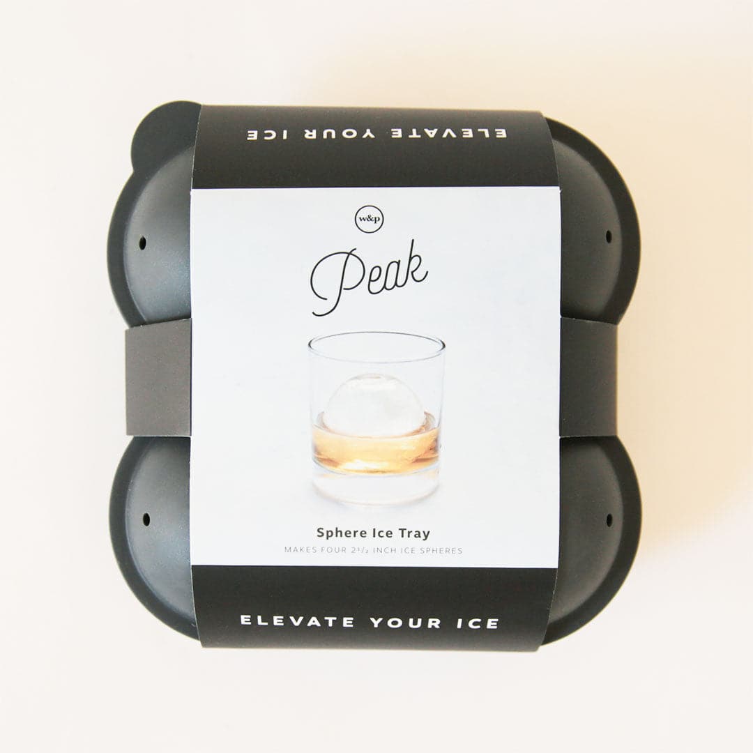 On a cream background is a charcoal silicone ice cube mold with a label on the outside that has a photo of the sphere shaped ice cube inside a glass with text that reads, &quot;Peak Elevate Your Ice&quot;.