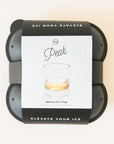 On a cream background is a charcoal silicone ice cube mold with a label on the outside that has a photo of the sphere shaped ice cube inside a glass with text that reads, "Peak Elevate Your Ice".