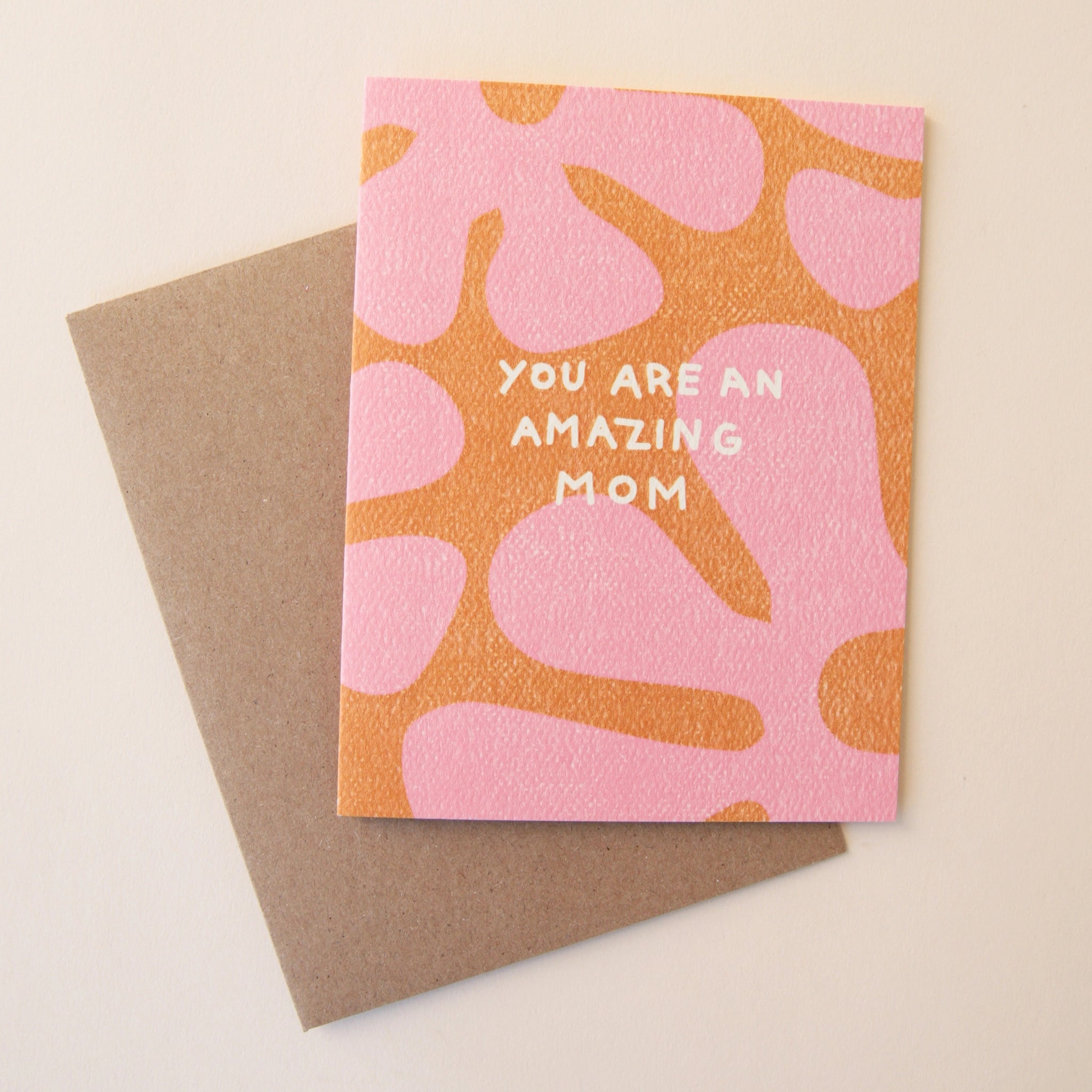 An orange card with a large pink floral print all over the front along with white text in the center that reads, "You Are An Amazing Mom" and a coordinating kraft brown envelope.