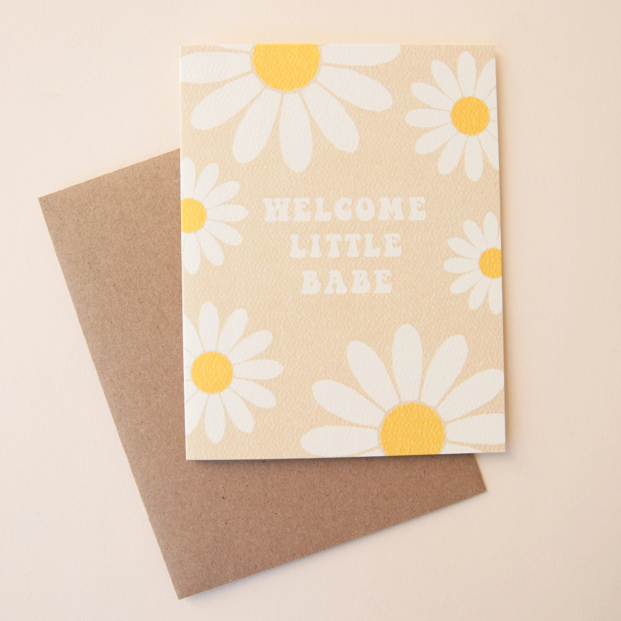 A tan card with white and yellow daisies all over the edges along with groovy text in the center that reads, &quot;Welcome Little Babe&quot; along with a Kraft brown envelope.