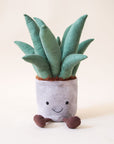 A stuffed animal aloe vera plant with a grey pot, two brown legs and a light green spiky aloe leaves along with a smiling face on the grey planter area.
