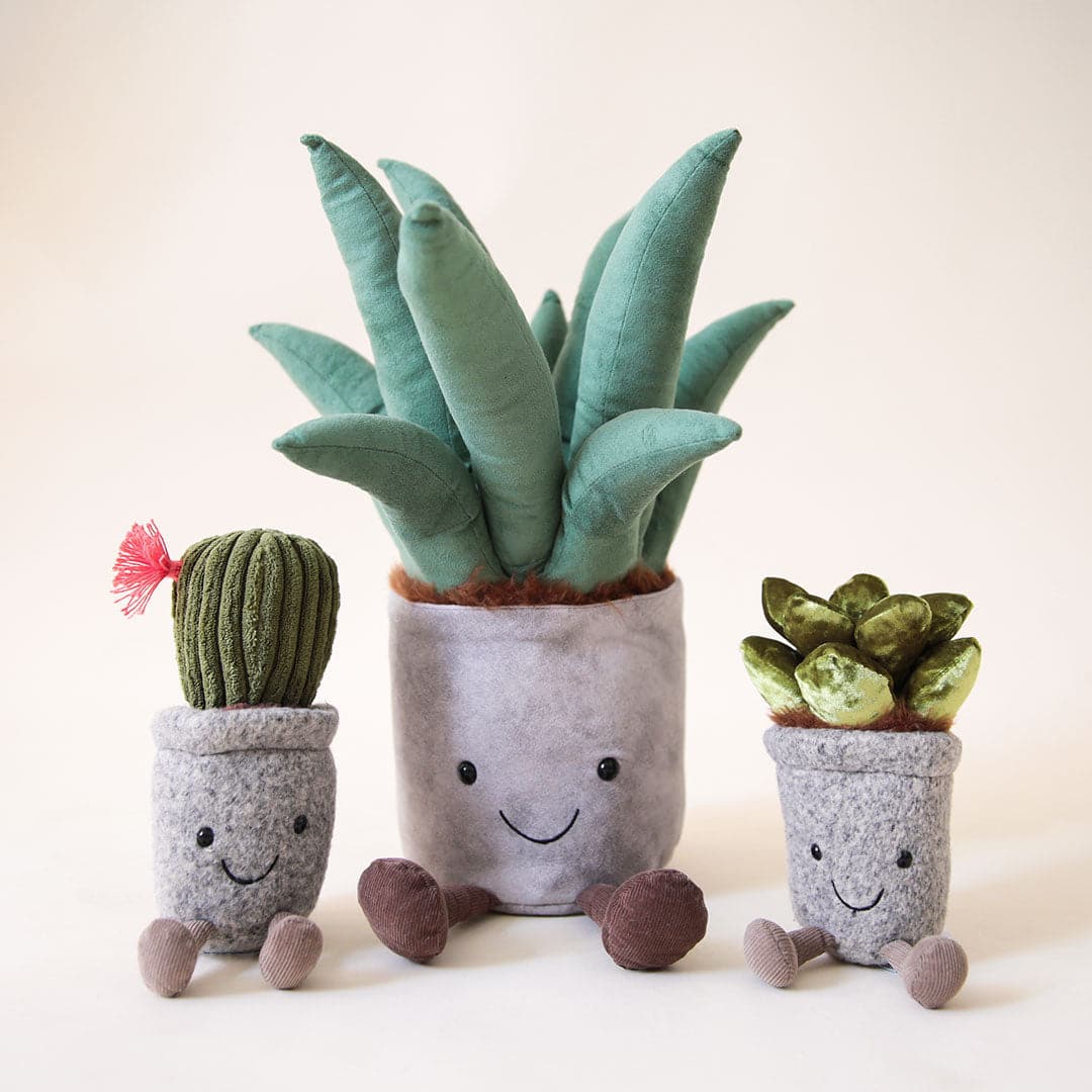 A stuffed animal aloe vera plant with a grey pot, two brown legs and a light green spiky aloe leaves along with a smiling face on the grey planter area photographed with two other cacti stuffed toys.
