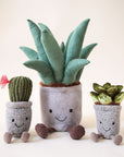 A stuffed animal aloe vera plant with a grey pot, two brown legs and a light green spiky aloe leaves along with a smiling face on the grey planter area photographed with two other cacti stuffed toys.