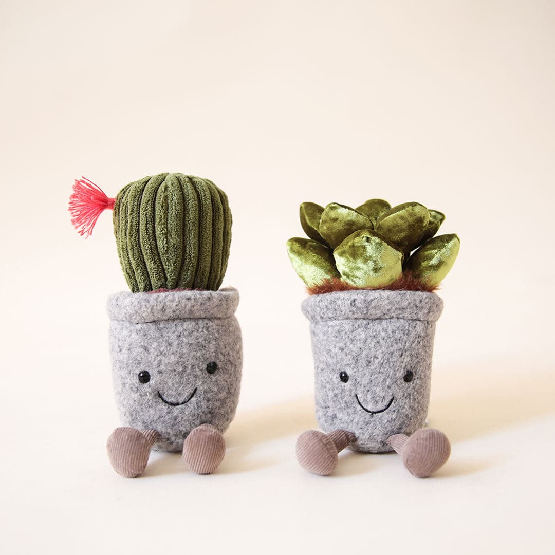 In front of a peachy background is a succulent stuffed animal. It is a gray pot with a black smiley face on the pot. It has two brown legs at the bottom of the pot. Inside the pot is brown with a shiny green succulent on top. The succulent has two layers of green leaves. To the left is another succulent stuffed animal. It is exactly like the one on the right but on top is a dark green cactus with a hot pink flower on the left side.