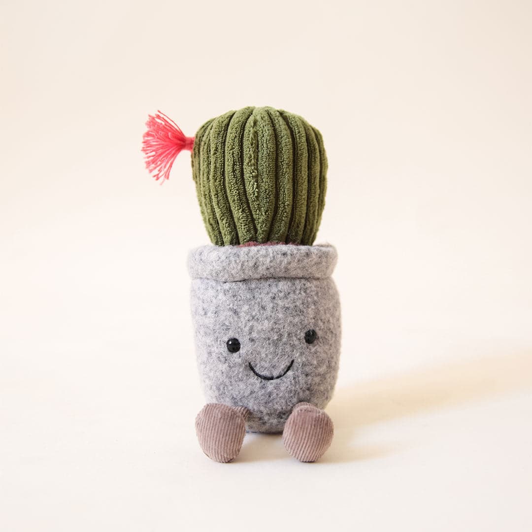 In front of a peachy background is a succulent stuffed animal. It is a gray pot with a black smiley face on the front and two brown legs on the bottom. On top of the pot is a dark green cactus with a hot pink flower on the left side.
