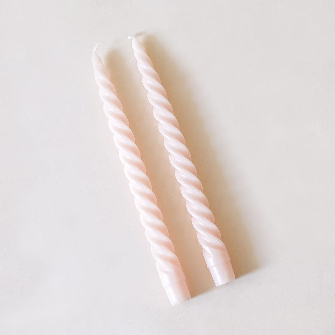 Pair of two blush pink, twisted taper candles with white cotton wicks lay beside each other against a white background.