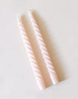 Pair of two blush pink, twisted taper candles with white cotton wicks lay beside each other against a white background.