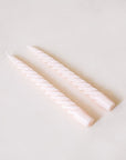 Pair of two blush pink, twisted taper candles with white cotton wicks lay beside each other against a white background.