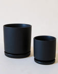 On a cream background is two different sized black ceramic pots with removable trays for watering.