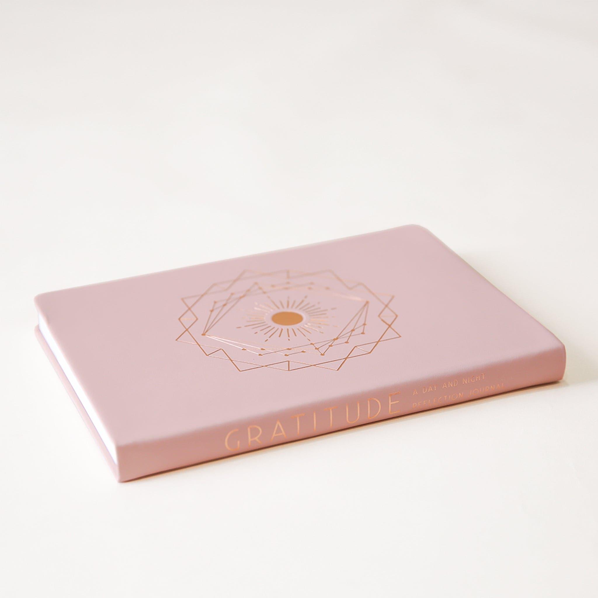 On a cream background is a light pink journal with a geometric gold design in the center and laying on its side in this photo showing the spine of the journal that reads, &quot;Gratitude&quot; in gold letters. 