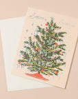 A cream colored card sits against a light peach background. On the front of the card there is a drawing of a dark green Christmas tree. There are yellow and red Christmas lights up and down the tree. There are green, red and pink ornaments spread out all over the tree. Silver tinsel is hanging on the end of each tree branch. At the bottom is a red tree skirt. At the top is a sliver star. Above the tree in silver script it reads ‘season's greetings.’ The card is accompanied by a white envelope.