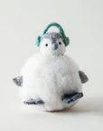 Sitting in front of a white background is a felt penguin ornament. The body is a white, round body. It has two little gray feet and two gray hands. On top is a little white and gray head with a gray nose, two black eyes and green ear muffs.
