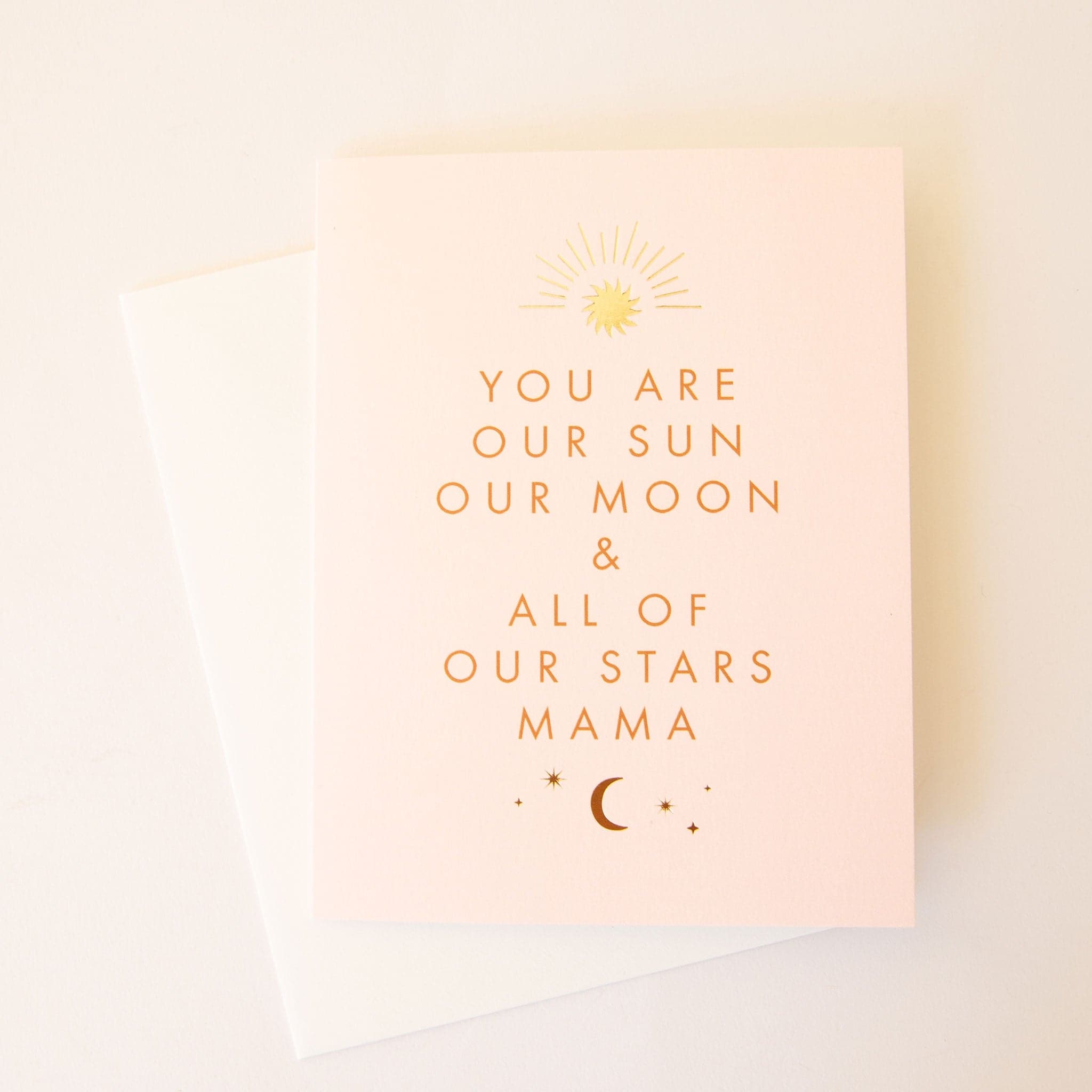 Pale pink card that reads 'you are our sun our moon & all of our stars mama' in orange capital lettering. A gold foil sun beams above the text and burnt orange crescent moon sits below. 