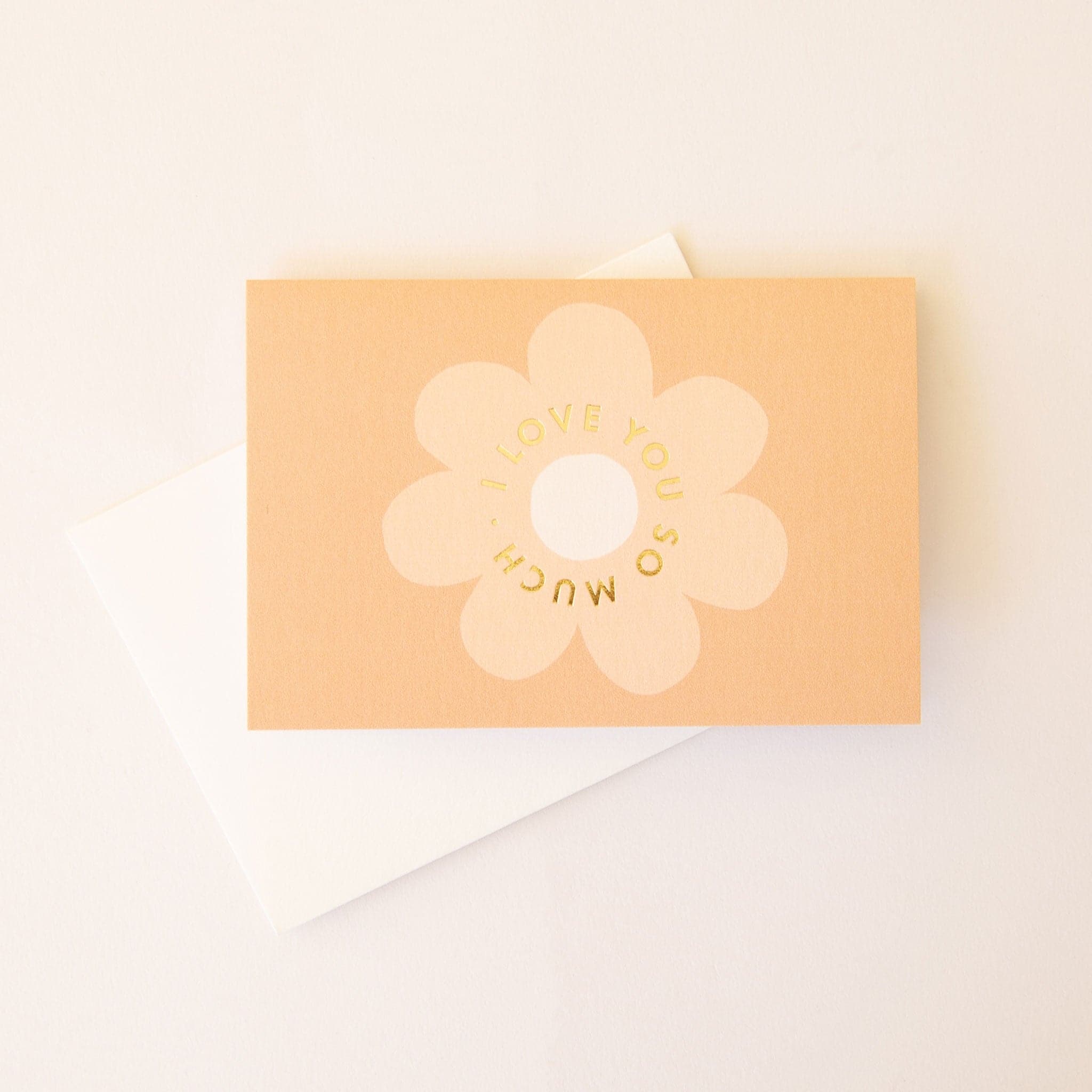 Pale orange horizontal card with a classic soft tan daisy flower in the center. The text &#39;I love you so much.&#39; in gold foil curves within the flower. The card is accompanied by a solid white envelope. 