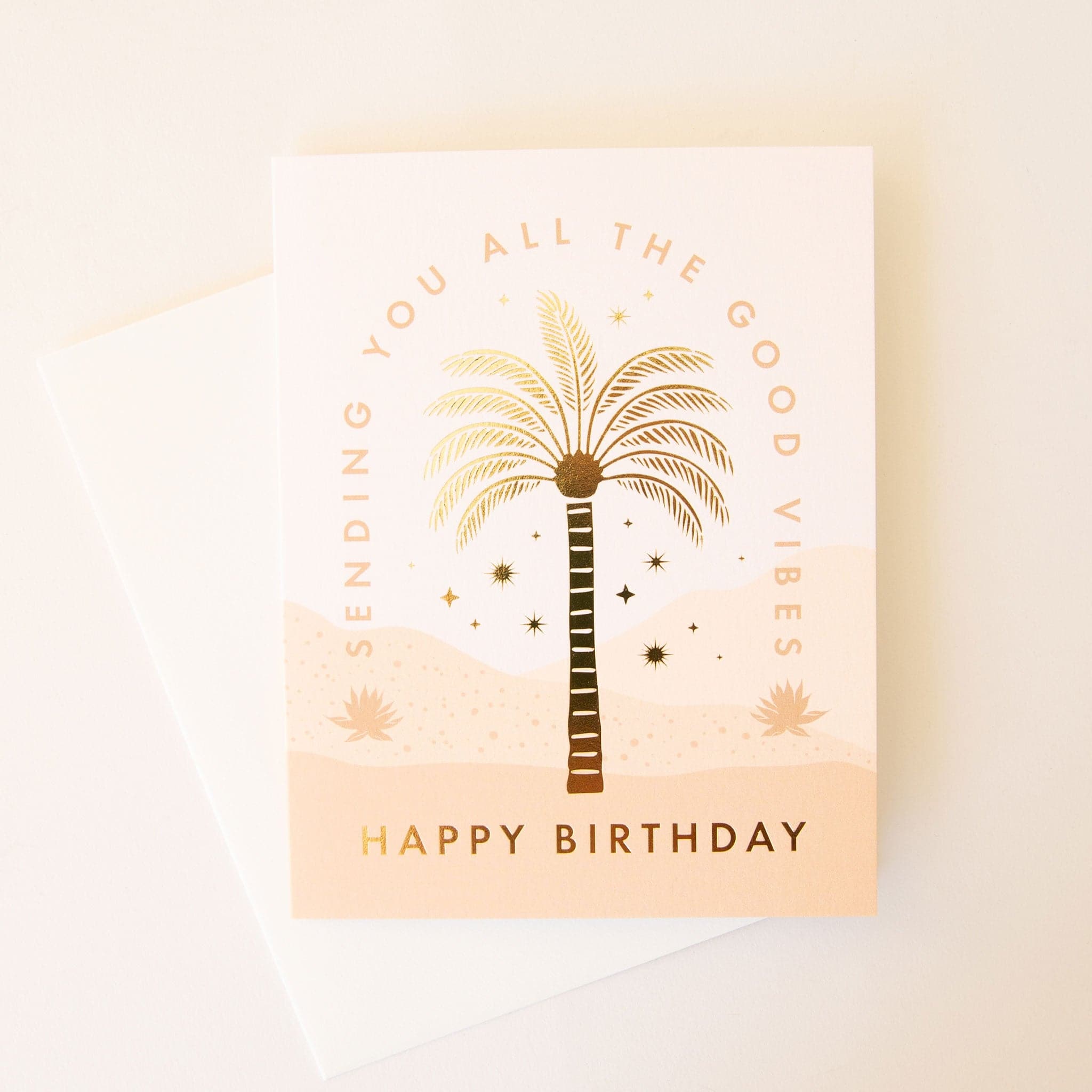 Warm toned birthday card complete with a desert scene. The text 'Sending you all the good vibes' curves around a gold foil palm tree and desert stars. Below the scene reads 'Happy Birthday' in capital gold foil lettering. The card is accompanied by a solid white envelope. 