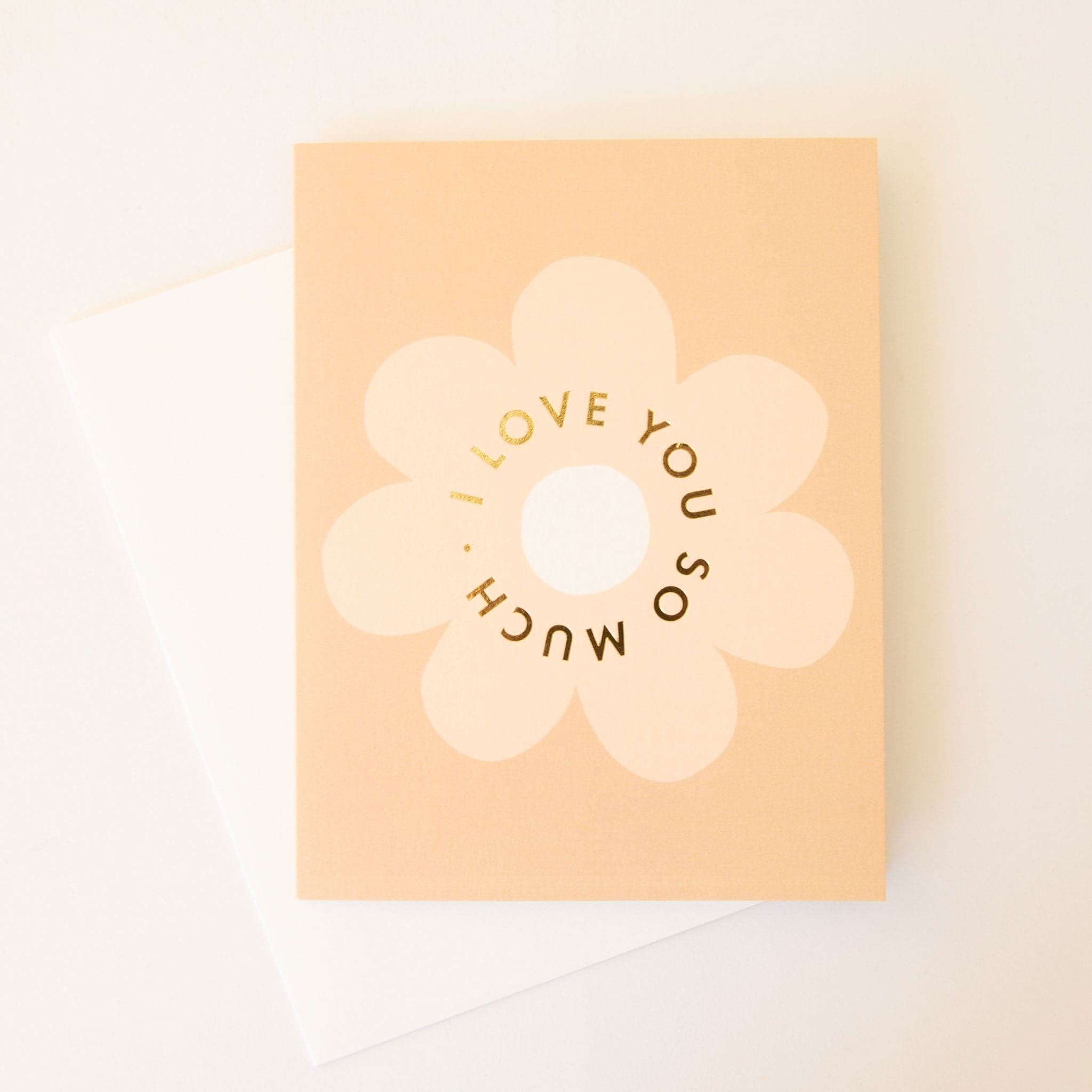 Pale orange card with a classic soft tan daisy flower in the center. The text 'I love you so much.' in gold foil curves within the flower. The card is accompanied by a solid white envelope. 