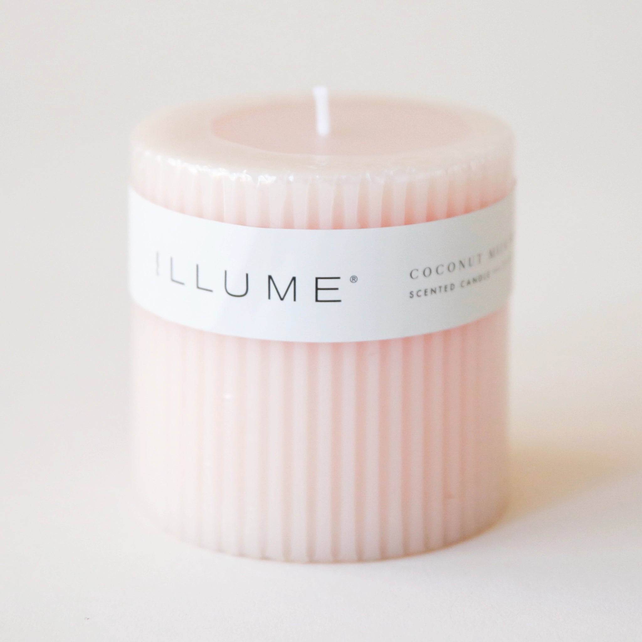A light pink pilar candle with a ribbed detail and a thin white label wrapped around that reads, &quot;Coconut Milk Mango Scented Candle&quot;.