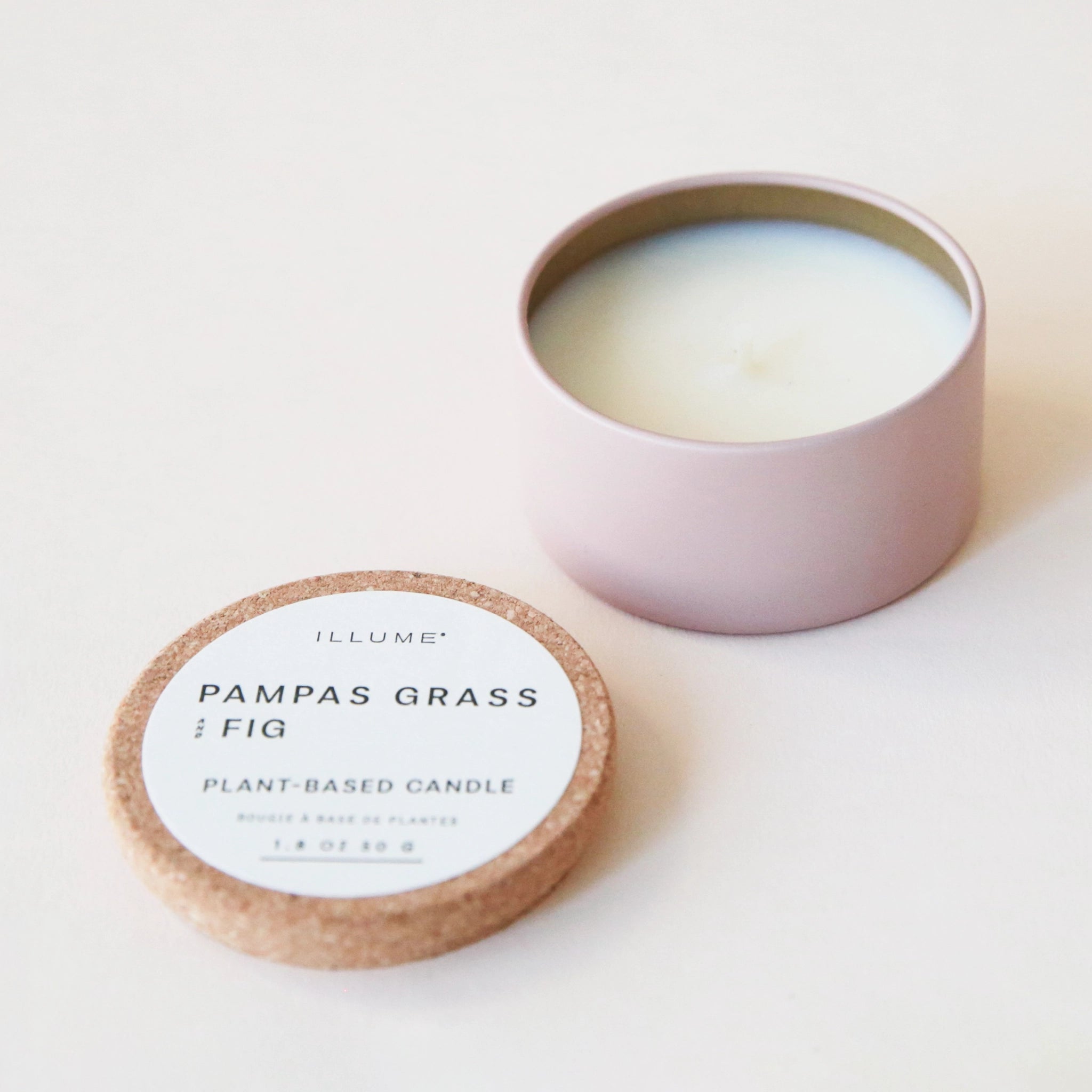 A pink tin with a white wax candle inside and a natural colored  cork lid with a white label that reads &quot;Pampas Grass &amp; Fig Plant Based Candle&quot; in black letters.