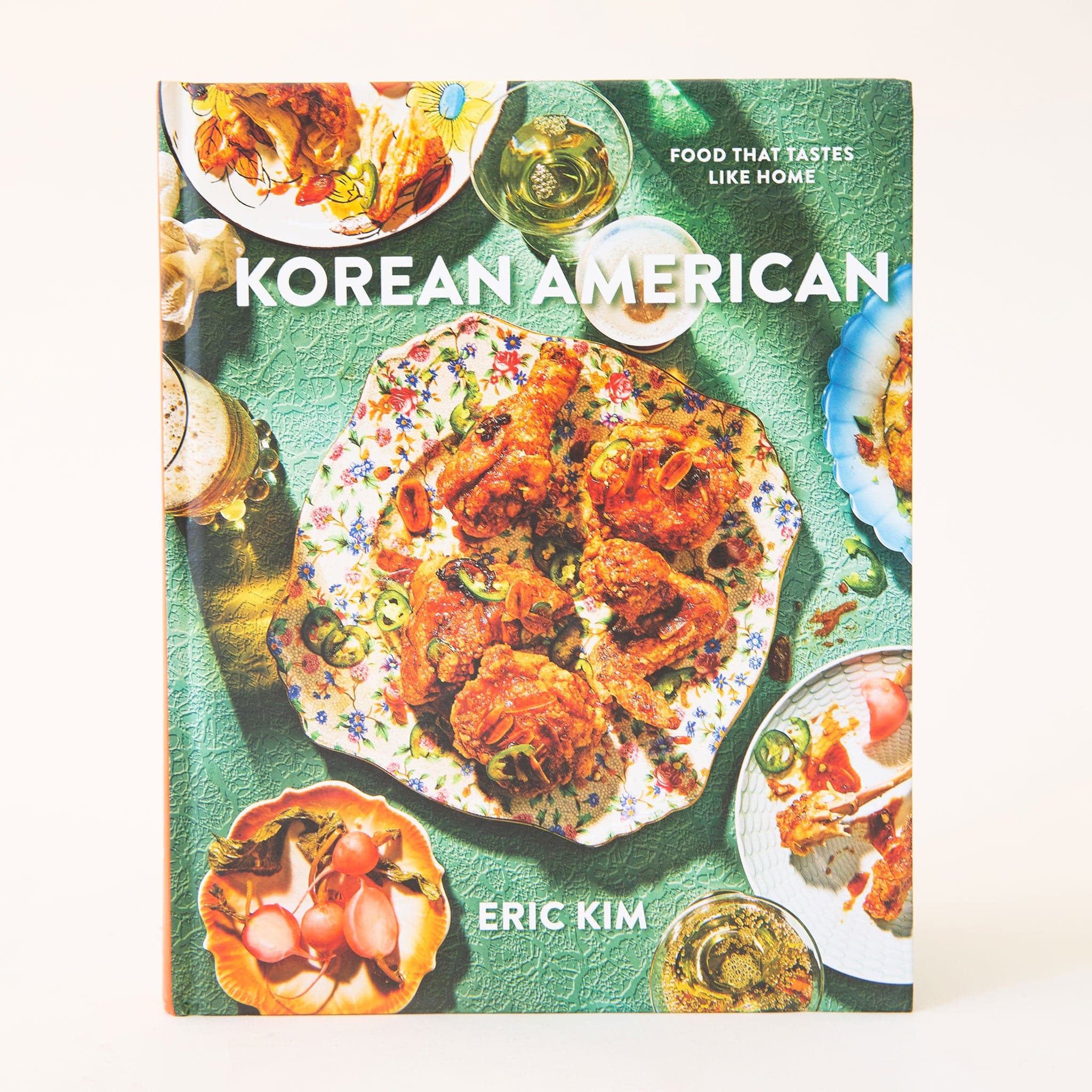 Hard cover cookbook titled &#39;Korean American&#39; in white bold lettering. The cover is filled with colorful dishes of beautifully curated meals. The plates lay against a sea green textured background. 