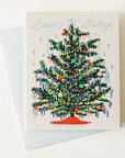 A cream colored card sits against a white background. On the front of the card there is a drawing of a dark green Christmas tree. There are yellow and red Christmas lights up and down the tree. There are green, red and pink ornaments spread out all over the tree. Silver tinsel is hanging on the end of each tree branch. At the bottom is a red tree skirt. At the top is a sliver star. Above the tree in silver script it reads ‘season's greetings.’ The card is accompanied by a white envelope.