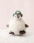 Sitting on a bed of fake snow and in front of a pink background is a felt penguin ornament. The body is a white, round body. It has two little gray feet and two gray hands. On top is a little white and gray head with a gray nose, two black eyes and green ear muffs.