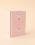 A small pink book that says, "Gratitude Meditations & Inspirations" in light pink on the front cover as well as s geometric illustration above and below the text.