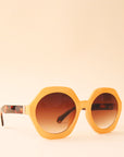 Mustard yellow circular frame sunglasses with a brown lens and tortoise arms photographed in front of a peachy background.