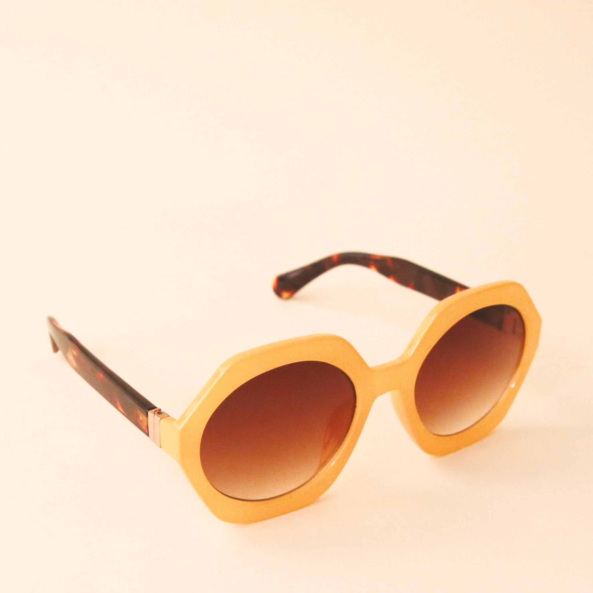 Mustard yellow circular frame sunglasses with a brown lens and tortoise arms photographed in front of a peachy background.