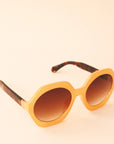 Mustard yellow circular frame sunglasses with a brown lens and tortoise arms photographed in front of a peachy background.