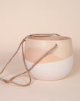 A ceramic circle planter with a wavy design featuring the top half as a neutral tan shade and the bottom half white along with three tan ropes tied together at the top in order to hang.