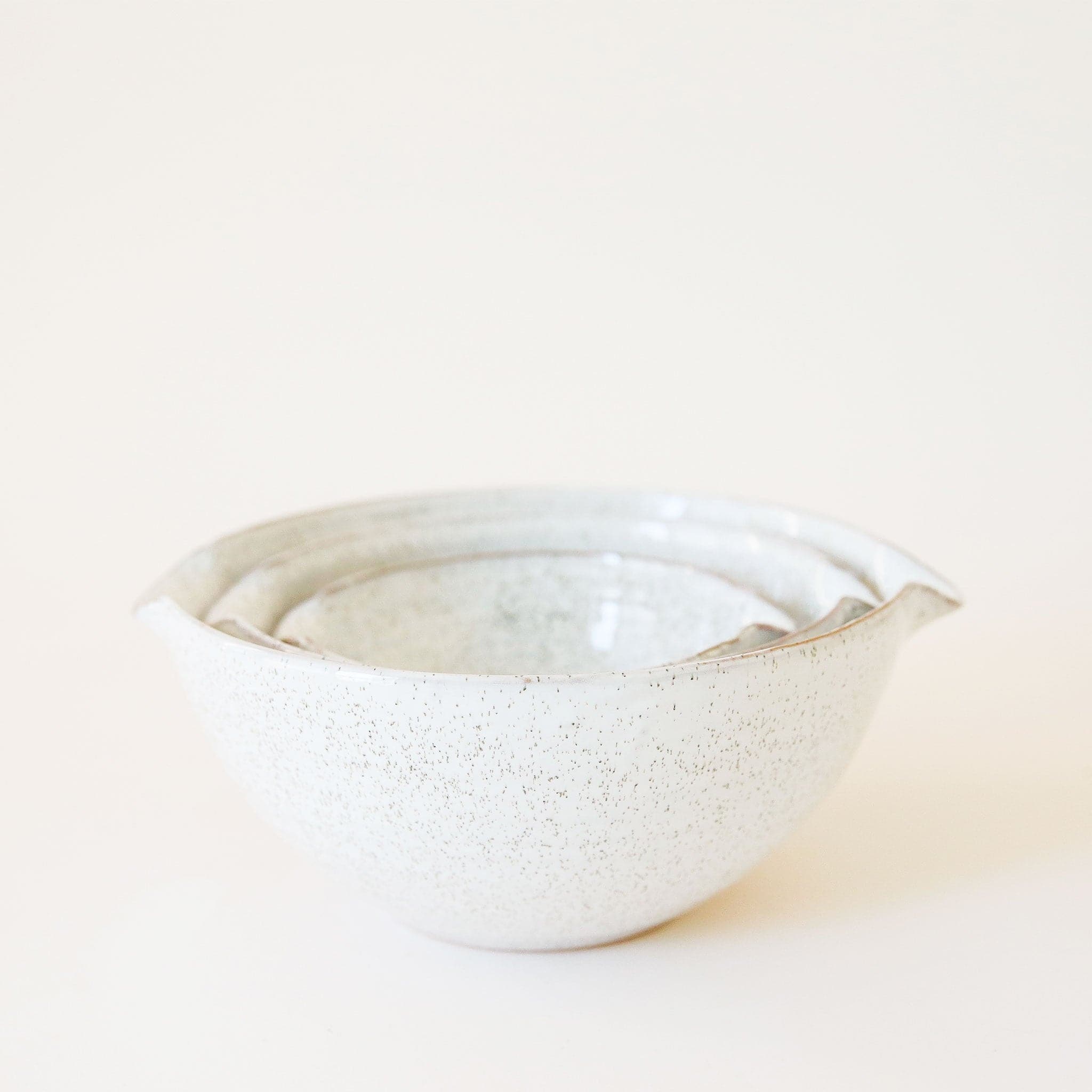 A set of three ceramic bowls nested inside one another. They feature a white base, glossy finish and a tan/brown speckled design. They are rounded bowls with a 'v' shaped design on two of the sides making them ideal for pouring if needed.