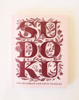 Pastel pink paperback puzzle book titled 'Sudoku' in large magenta capital lettering. The letters are accented with delightful floral detailing. Below the large lettering reads 'One hundred and fifty puzzles' towards the bottom of the cover.