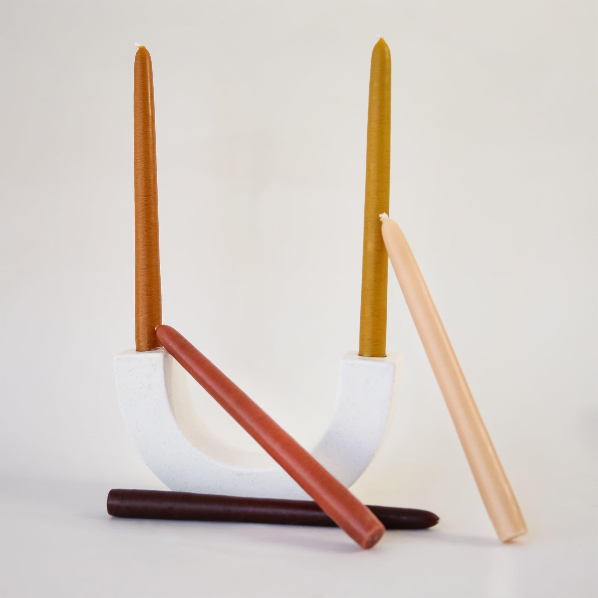 Two earth toned candles positioned on white 'U' shaped candle holder. Three candles in shades of peach and magenta lay against the candle holder.