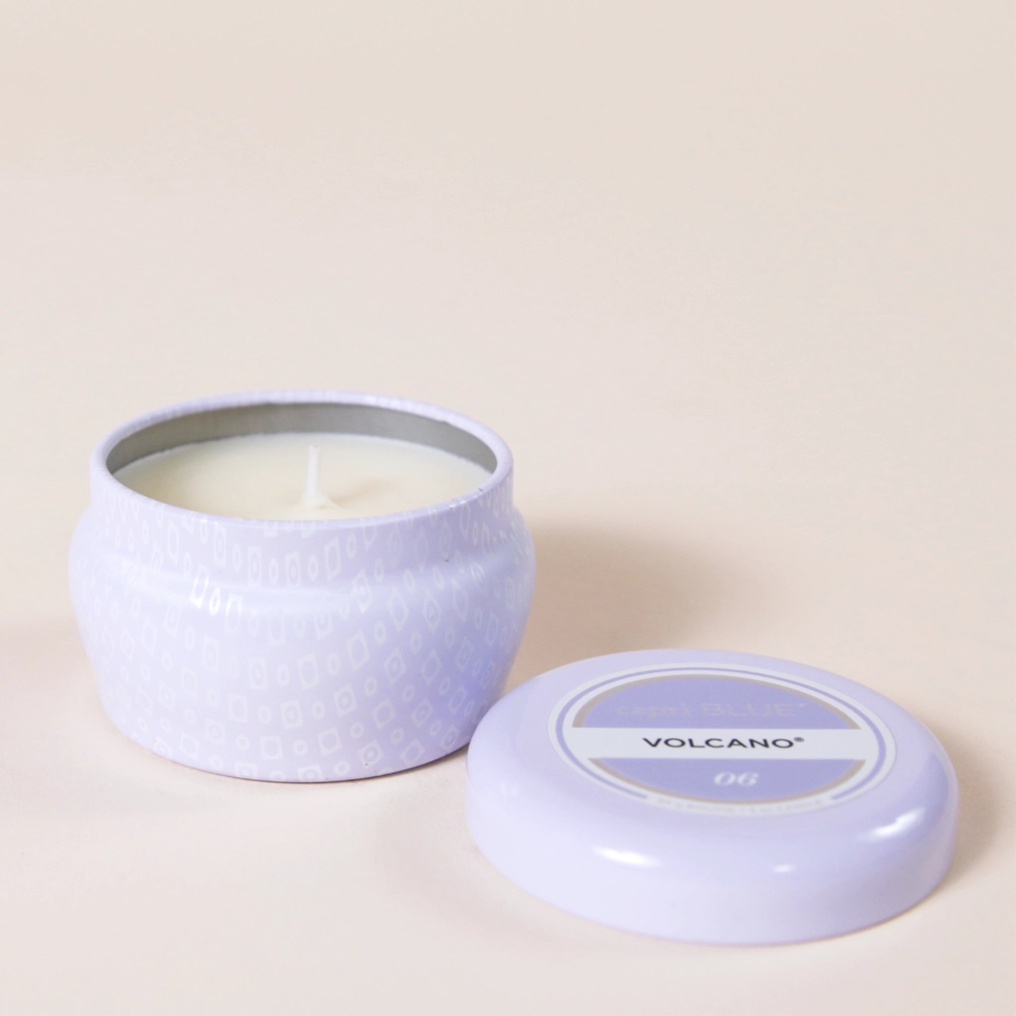 A small lavender tin candle with white wax and a white diamond repeating pattern on the outside of the tin. Comes with a matching purple lid that reads, "Capri Blue Volcano".