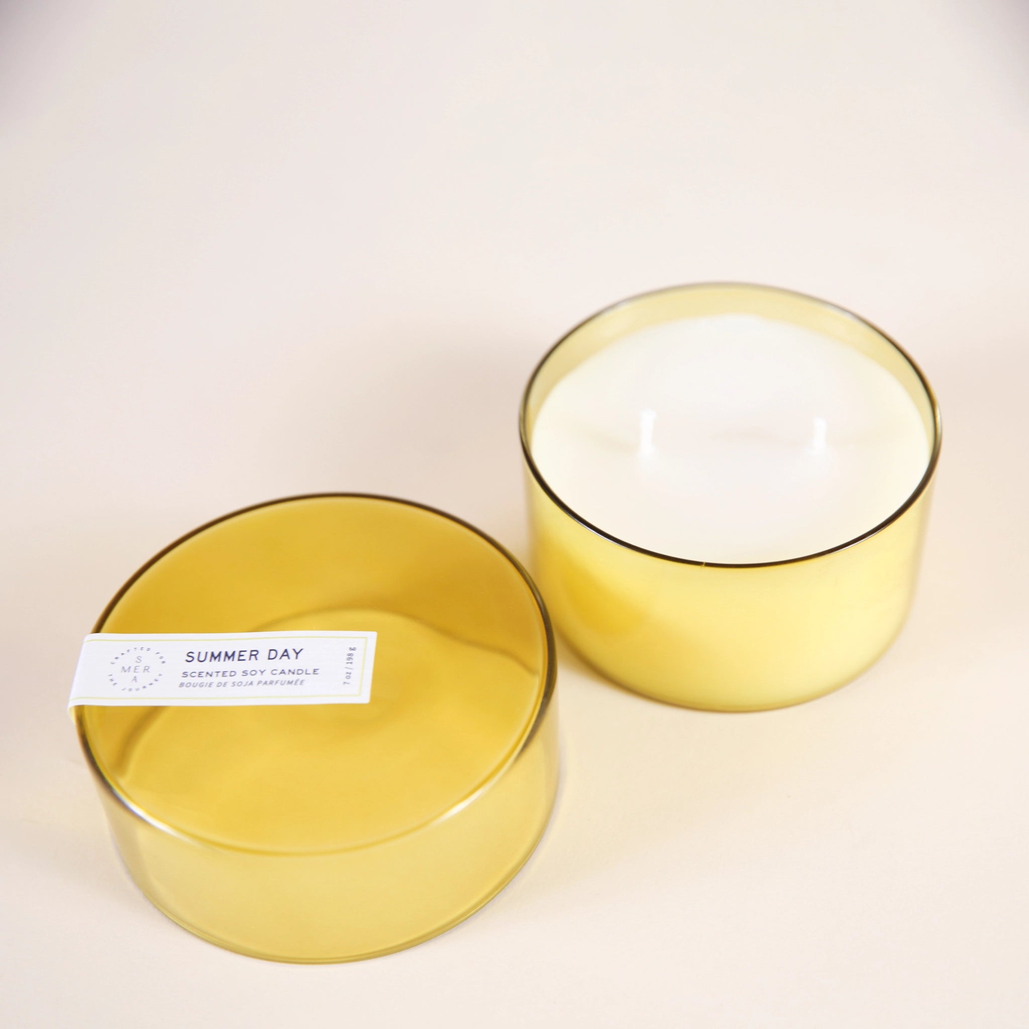 On a cream background is a yellow glass candle with a white double wick candle inside along with a coordinating yellow glass lid.