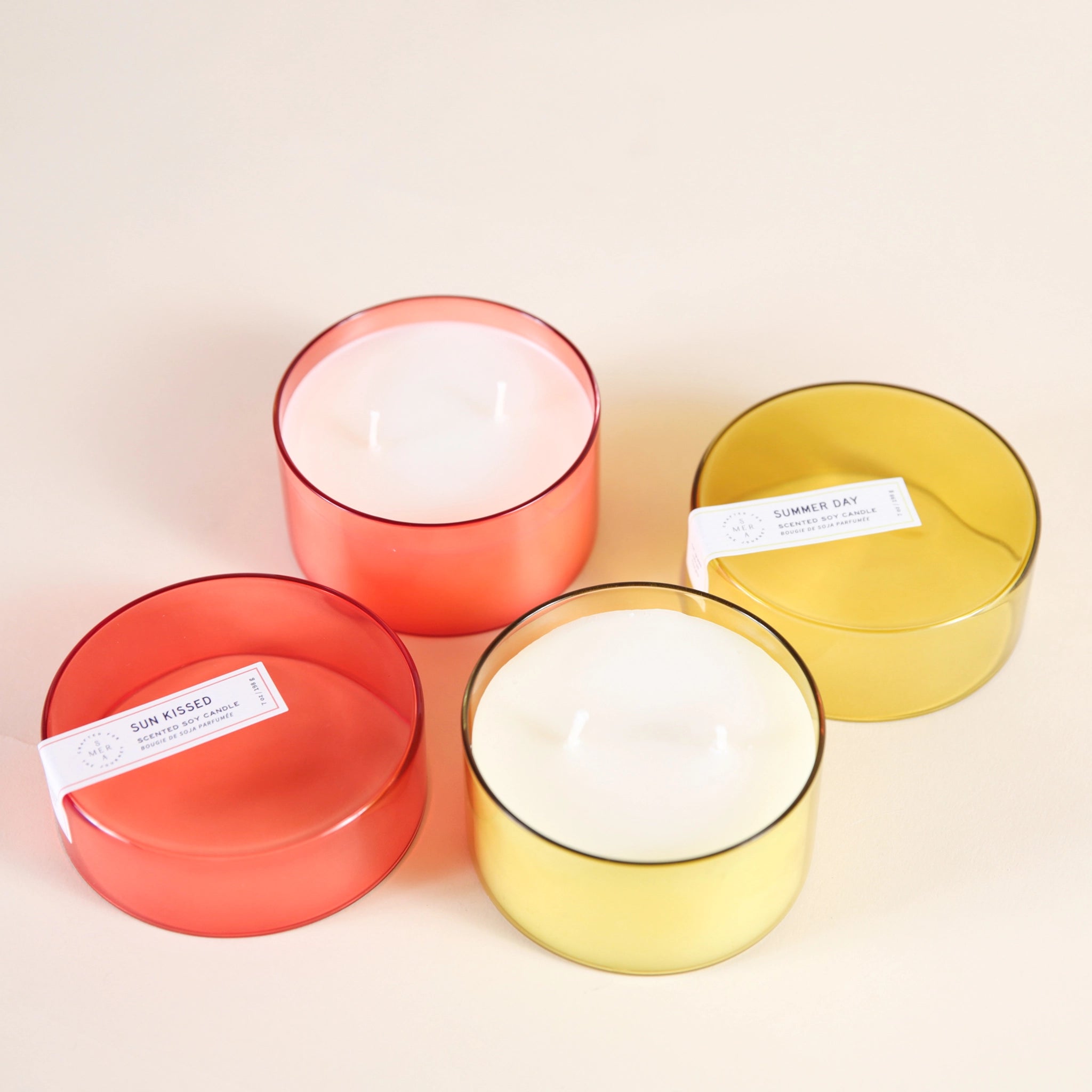 On a cream background is a yellow glass candle with a white double wick candle inside along with a coordinating yellow glass lid sitting next to the same candle but in a red color way. 