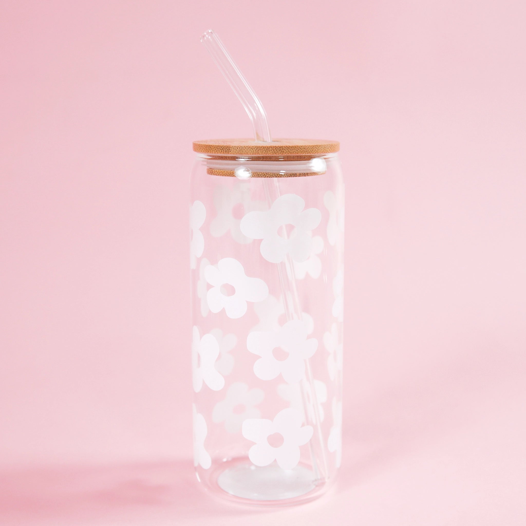 On a light pink background is a clear glass tumbler with a wood lid, a glass straw and white daisy designs all over.