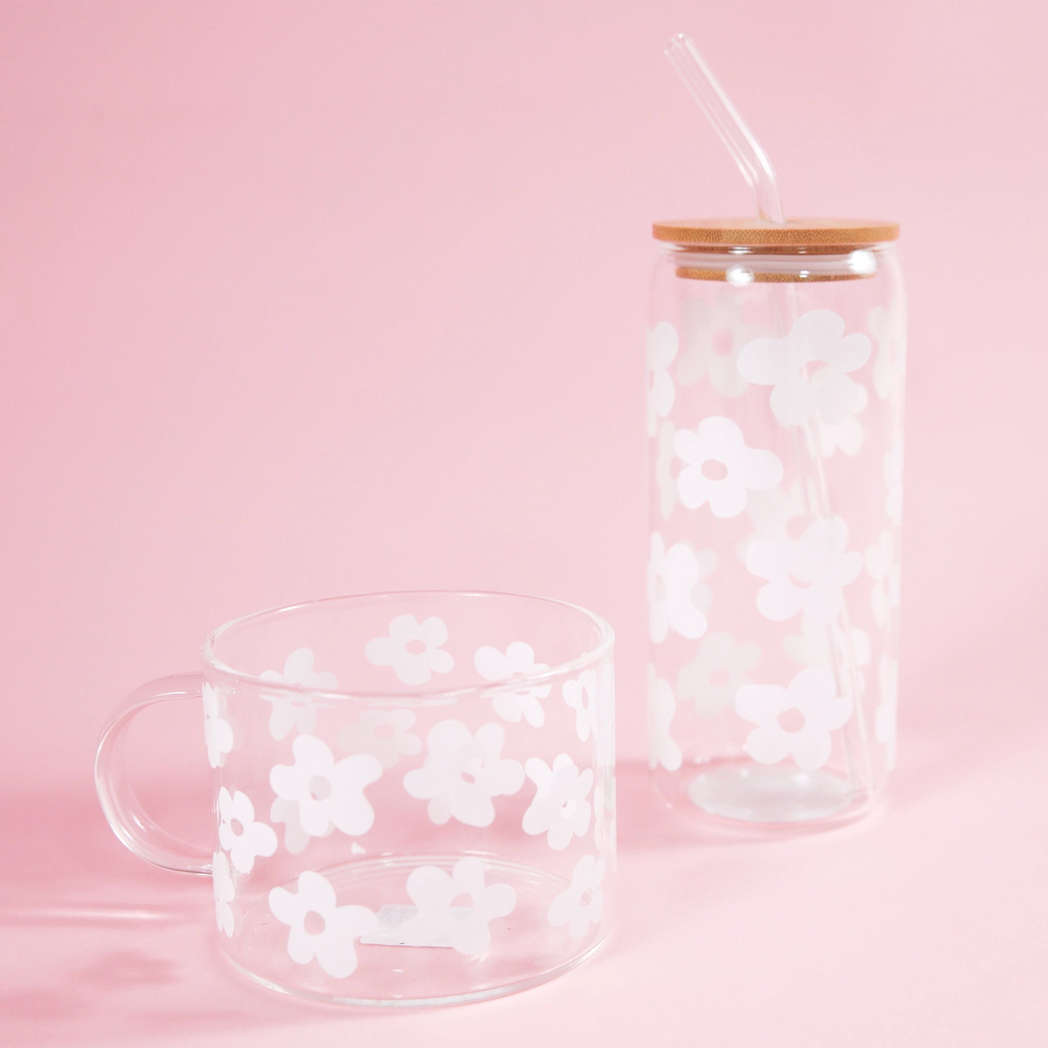 On a light pink background is a clear glass mug with a white daisy design all over photographed next to a taller glass tumbler with a wood lid and glass straw.