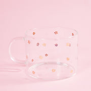On a light pink background is a clear glass mug with small gold daisy design all over.