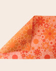 Sheet of wrapping paper filled with orange and pink flowers. The sheet is bent forward, revealing the other side of the wrapping paper. The back side is covered in the same floral print in oranges and yellows.