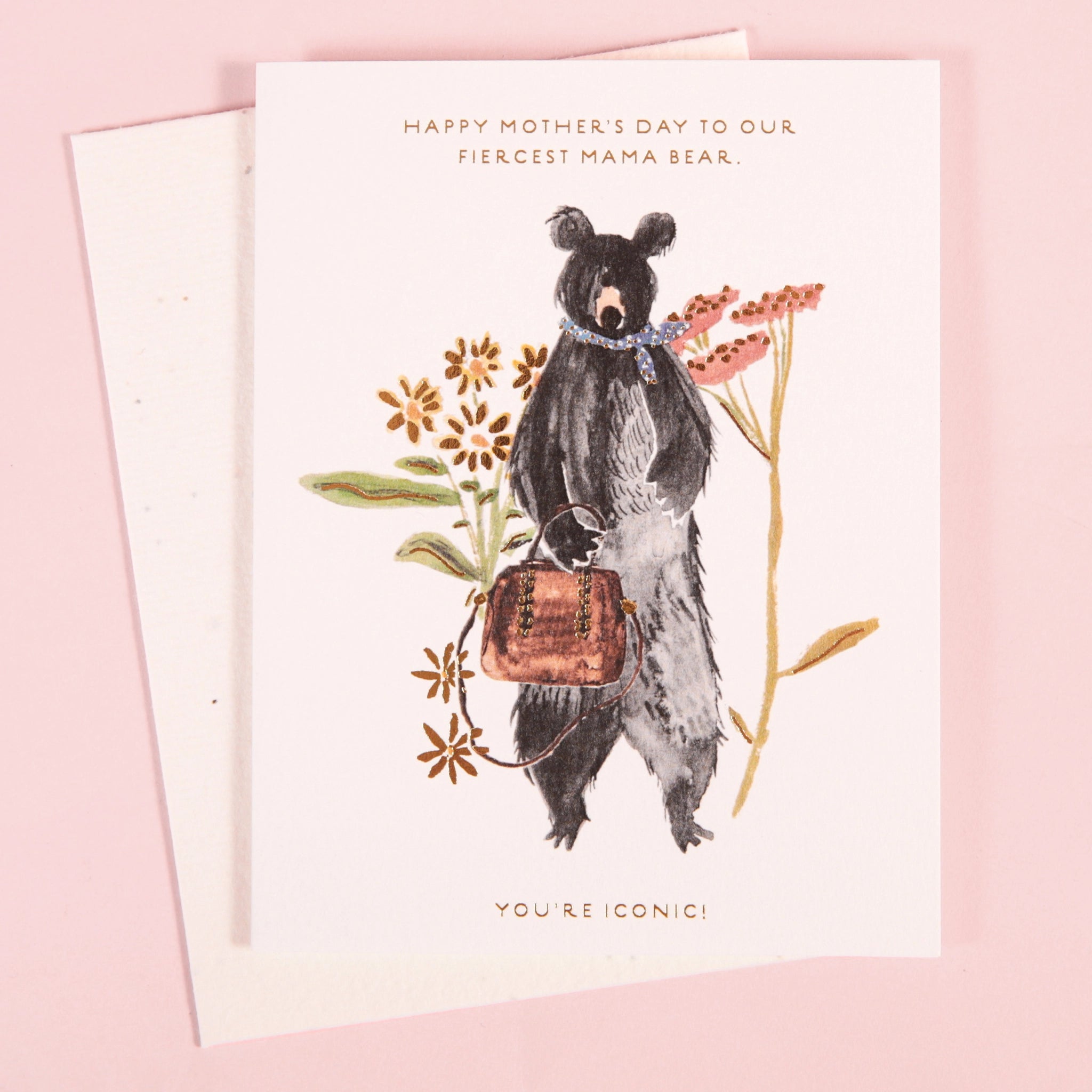 On a light pink background is a white card and envelope with an illustration of a black bear standing up on its hind legs while holding a brown purse in front of various flowers and text on the top that reads, &quot;Happy Mother&#39;s Day to our fiercest mama bear&quot;.