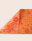 Sheet of wrapping paper filled with orange and yellow flower print. The sheet is bent forward, revealing the other side of the wrapping paper. The back side is covered in the same floral print in pink and oranges.