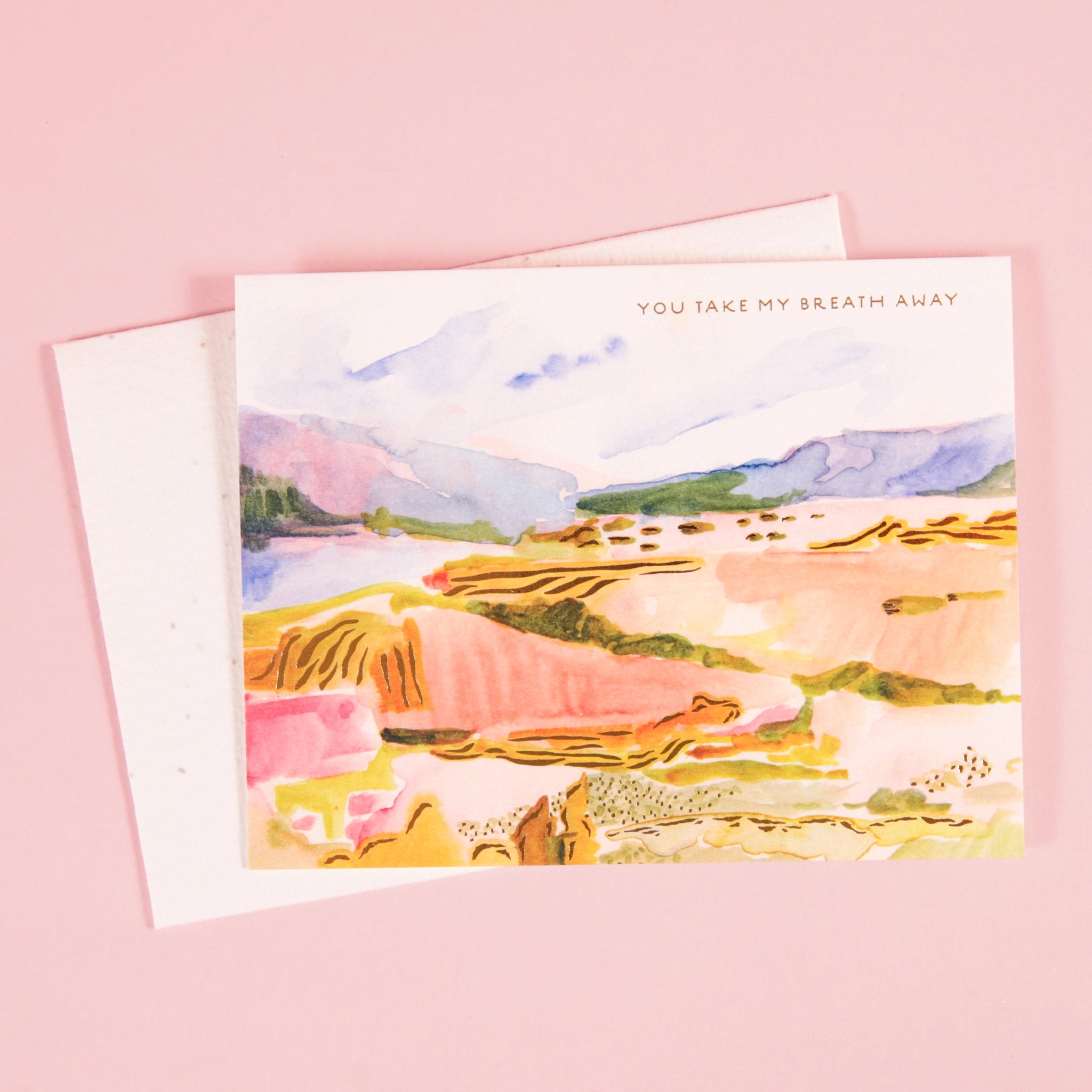 On a light pink background is a white envelope and card with a colorful illustration of a landscape and small text in the top right corner that reads, "You take my breath away".