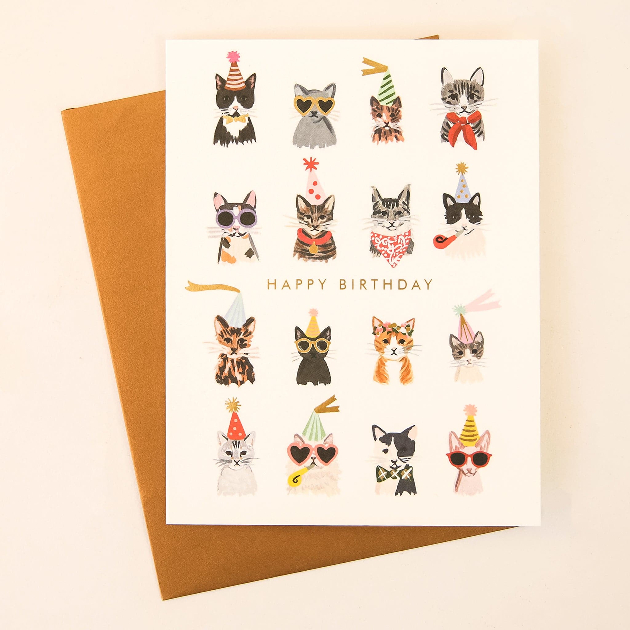 On a cream background is a photograph of a white card and a gold envelope. On the card is 16 various breeds of cats in all different colors wearing party hats, sunglasses and bowties along with gold text in the center that reads, &quot;Happy Birthday&quot;.
