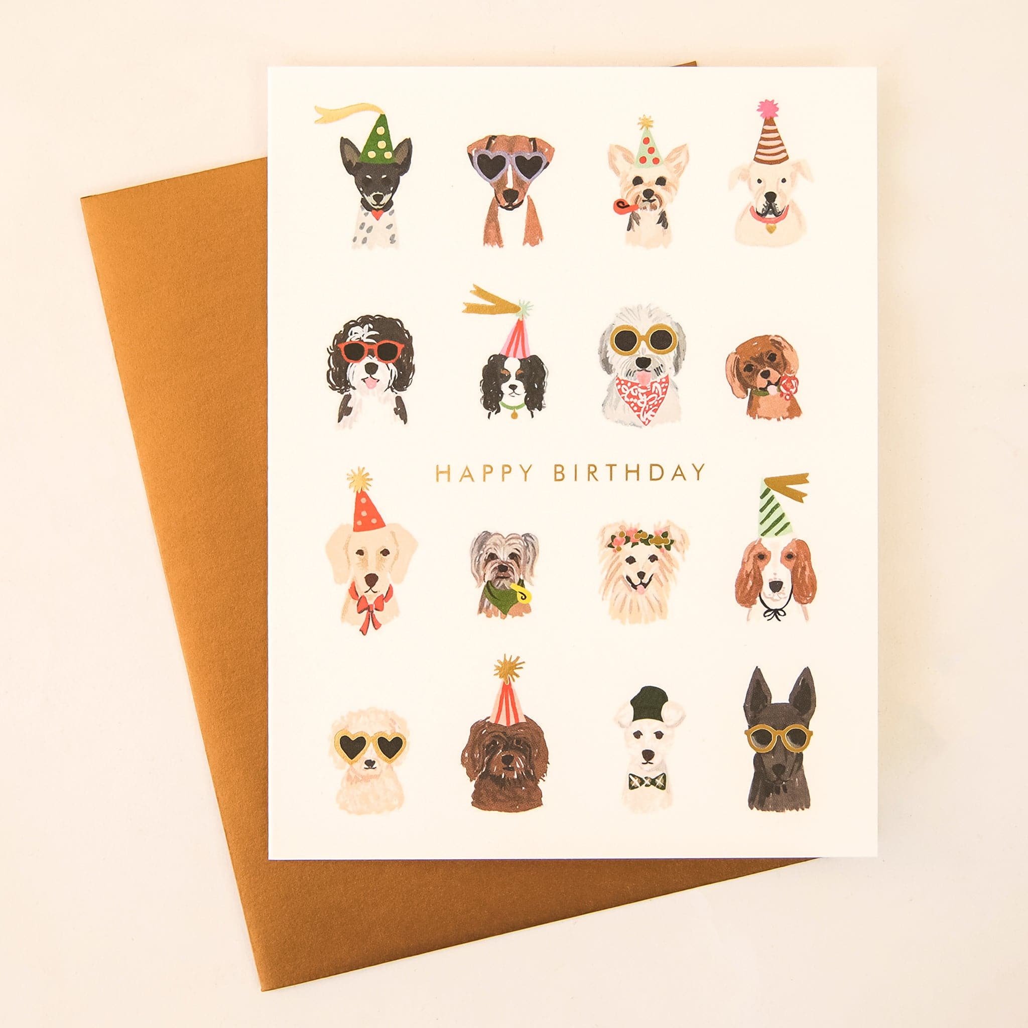 On a cream background is a gold envelope and a white card with various breeds of dogs in party attire and sunglasses as well as text in the center that reads, "Happy Birthday" in gold letters. 