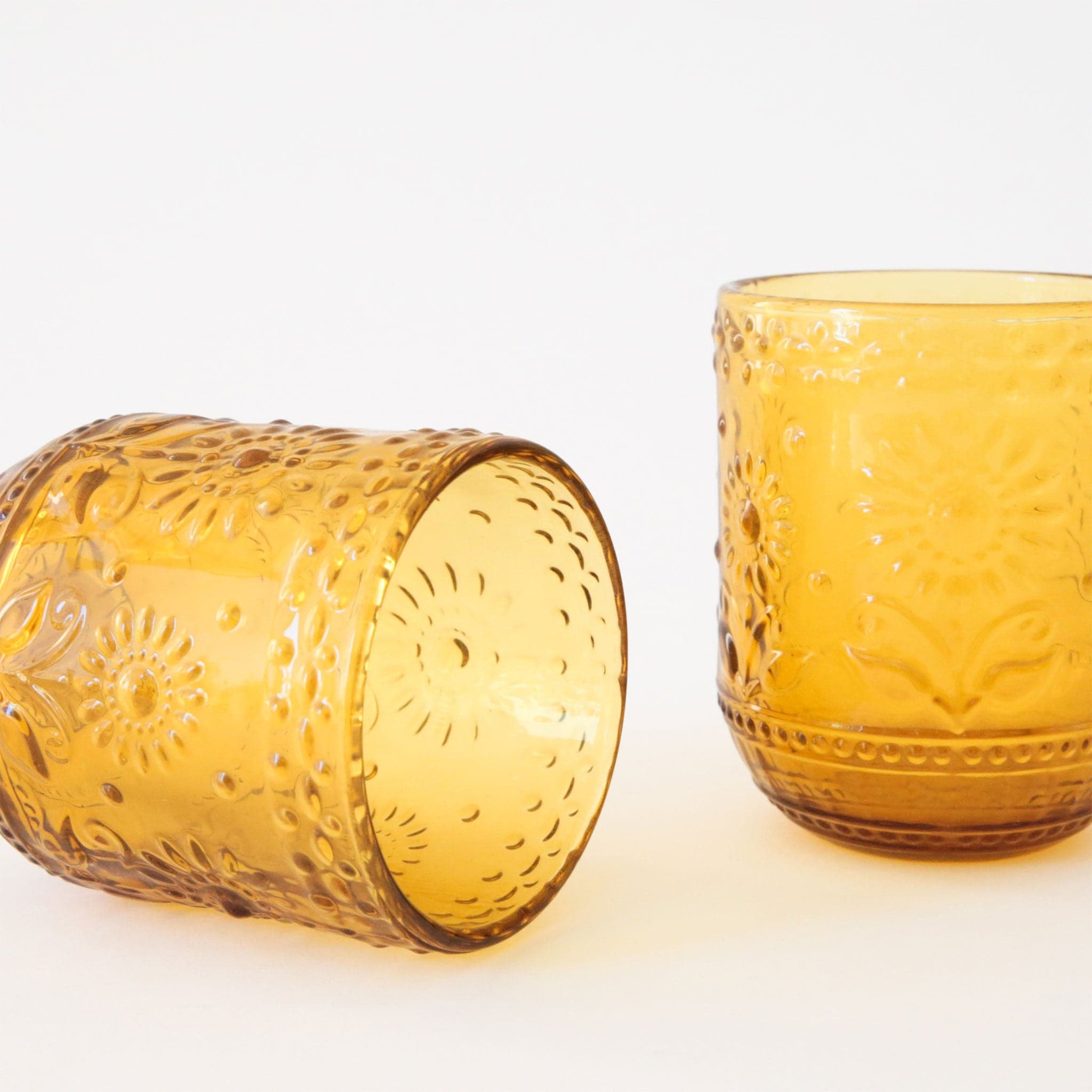 Two vintage inspired, embossed yellow drinking glasses with raised floral detailing. The glass to the left lays on its side and the glass to the right sits upright. 