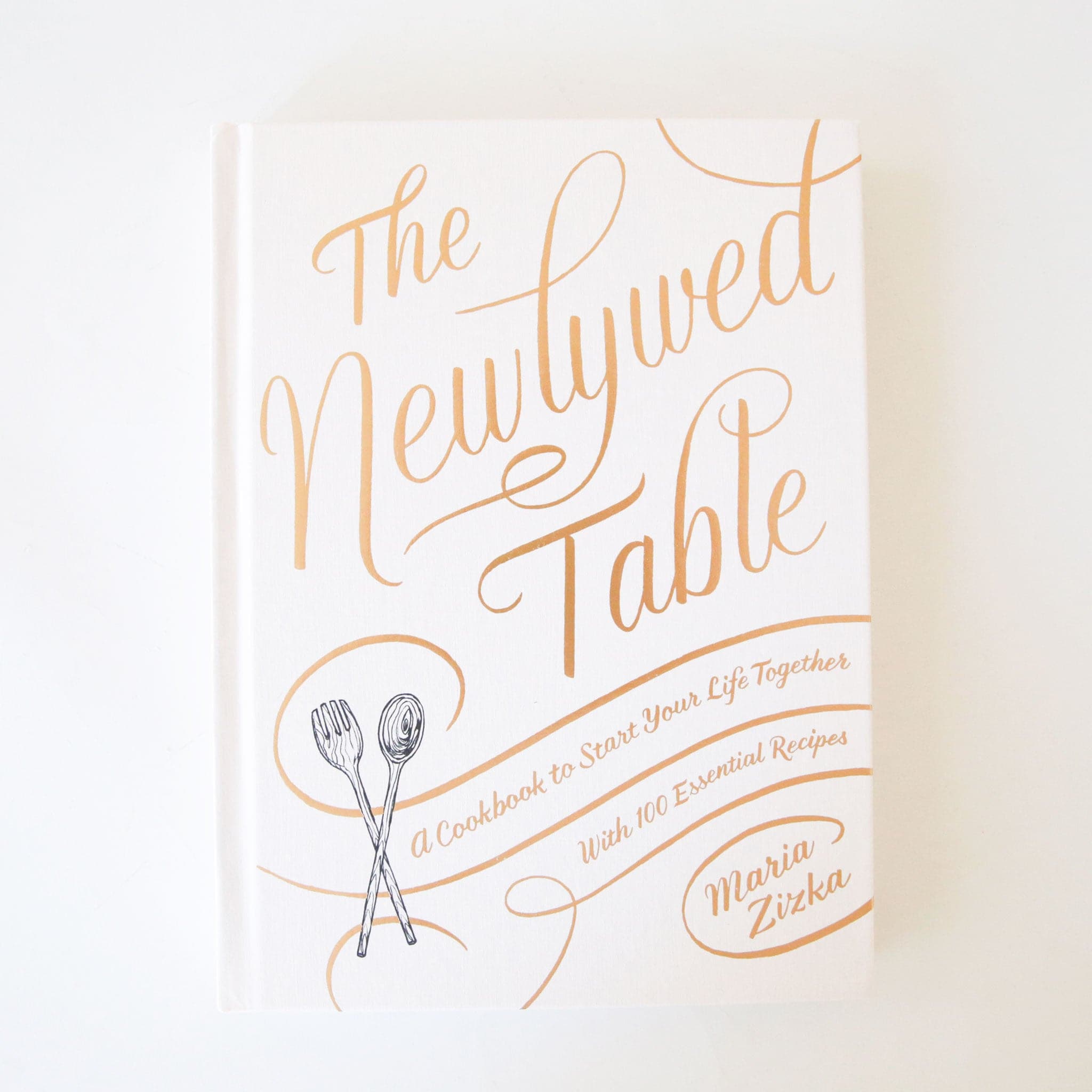 White hardcover book titled 'The newlywed table' in gold foil cursive lettering. Below reads 'a cookbook to start your life together with 100 essential recipes'. A black and white wooden spork and spoon sits in the bottom left corner. 