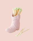 On a white background is a pink ceramic cowboy boot shaped match holder, photographed here with matches.